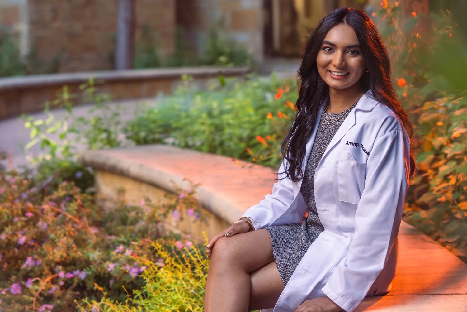 A young woman in a medical white coat and dress sits next to a garden near a limestone building, smiling.
