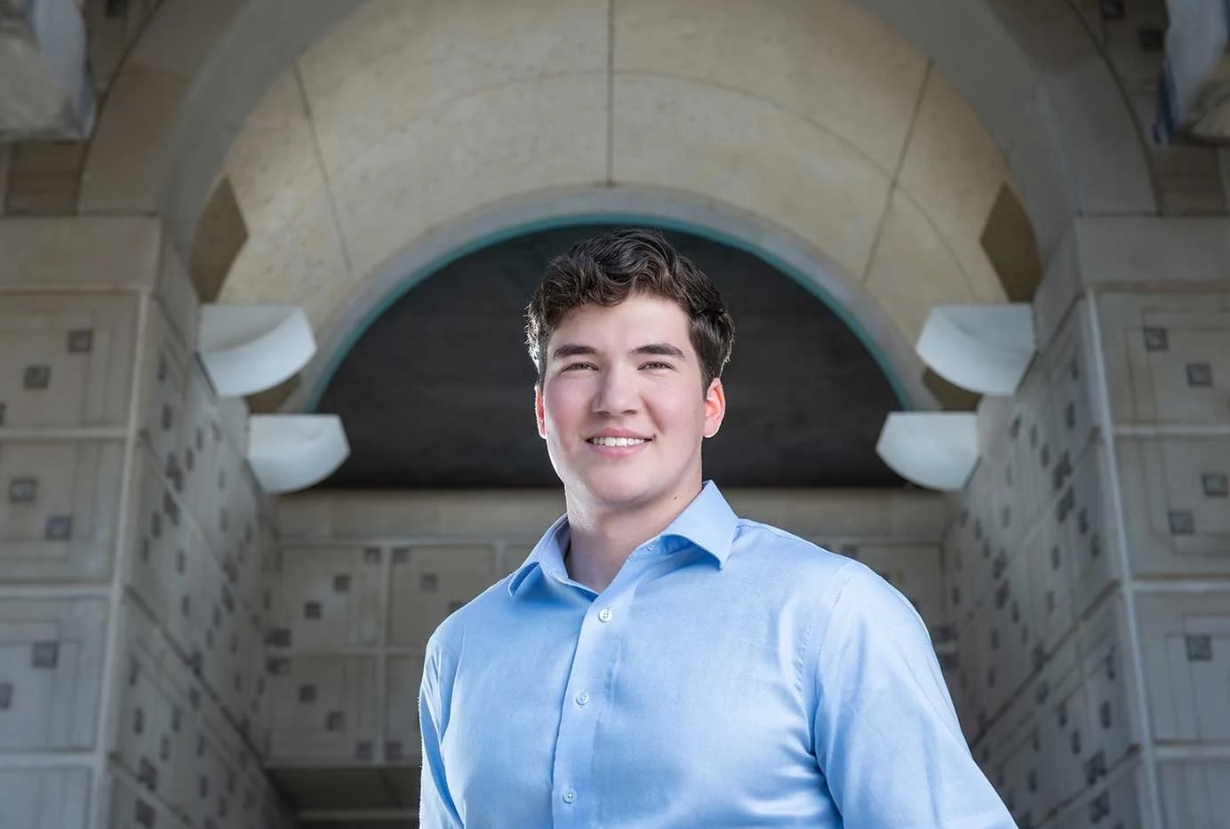 A young man in a collared shirt smiles while posing under an archway.